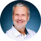 René Welter, ein Mann mittleren Alters mit kurzen grauen Haaren und Bart, lächelt. Er trägt ein weißes Hemd mit Kragen und posiert vor einem blauen Hintergrund.