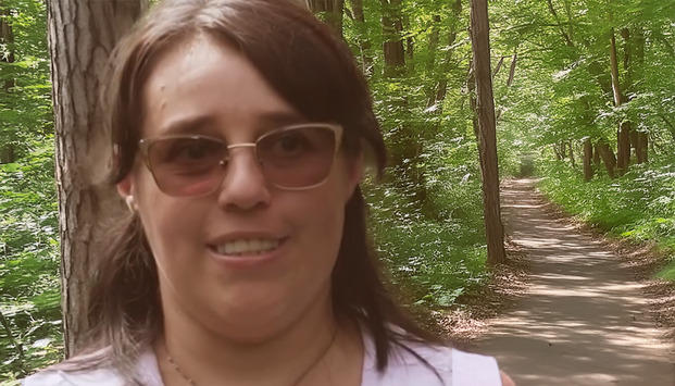 Im Vordergrund lächelt eine Frau mit braunen Haaren und Brille, die auf einem schattigen Waldweg steht, der von grünen Bäumen gesäumt ist.