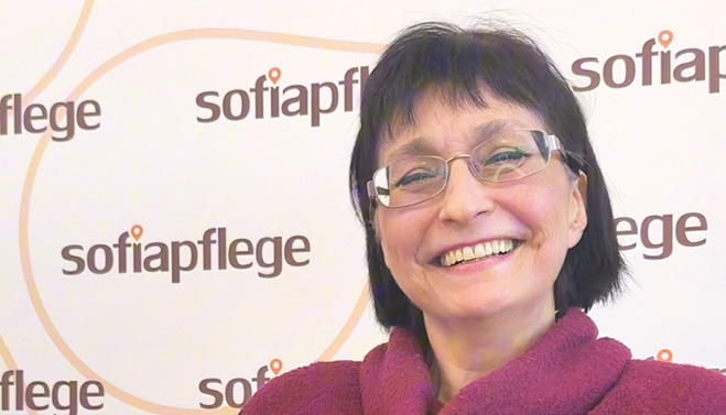 Eine lächelnde Frau mit kurzen dunklen Haaren und Brille steht vor einer Wand, auf der das Wort „sofiapflege“ wiederholt zu lesen ist.