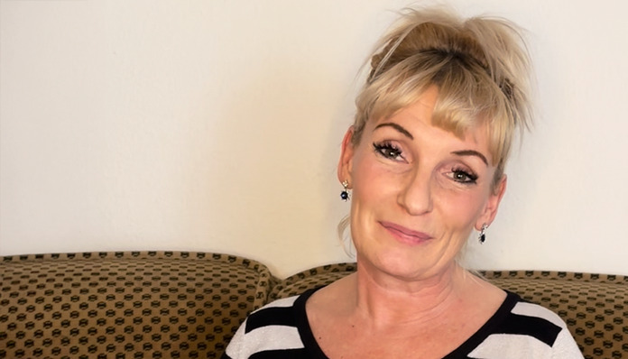 Eine Frau mit blonden, hochgesteckten Haaren sitzt auf einem gemusterten braunen Sofa und lächelt sanft. Sie trägt ein schwarz-weiß gestreiftes Oberteil und kleine Ohrringe. Im Hintergrund ist eine schlichte weiße Wand zu sehen.