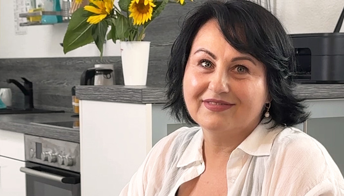Eine Frau mit dunklen Haaren und einem weißen Hemd sitzt in einer modernen Küche und lächelt. Im Hintergrund sind Sonnenblumen in einer weißen Vase zu sehen.