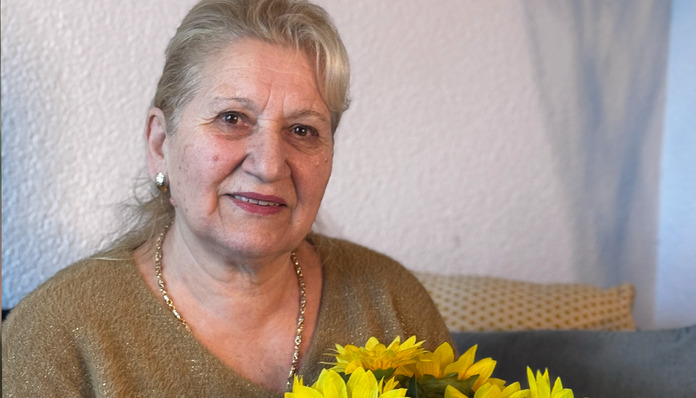 Eine ältere Frau mit blonden Haaren lächelt, während sie einen Strauß gelber Sonnenblumen in den Händen hält. Sie trägt einen goldenen Pullover und sitzt in einem Raum, im Hintergrund ist eine helle Wand zu sehen.