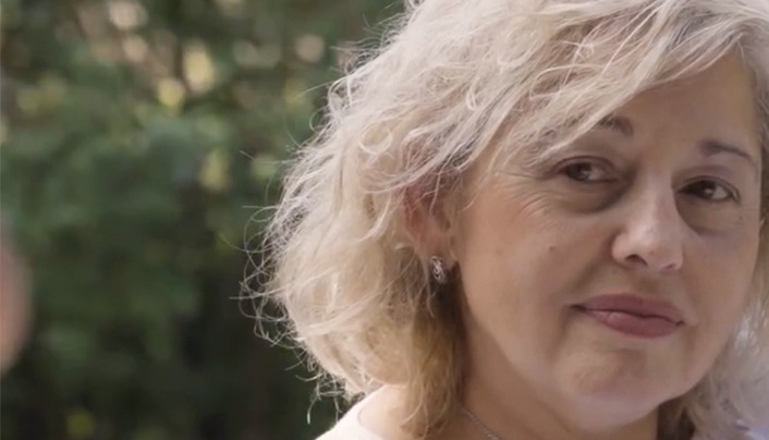 Eine Frau mit welligem blondem Haar blickt leicht von der Kamera weg und steht im Freien, während im unscharfen Hintergrund Grün zu sehen ist.