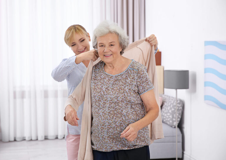 Eine junge Frau hilft einer älteren Dame in einem hellen, gemütlichen Wohnzimmer beim Anziehen einer beigefarbenen Strickjacke. Das Lächeln der älteren Dame spiegelt die warme und unterstützende Atmosphäre der 24-Stunden-Pflege wider.