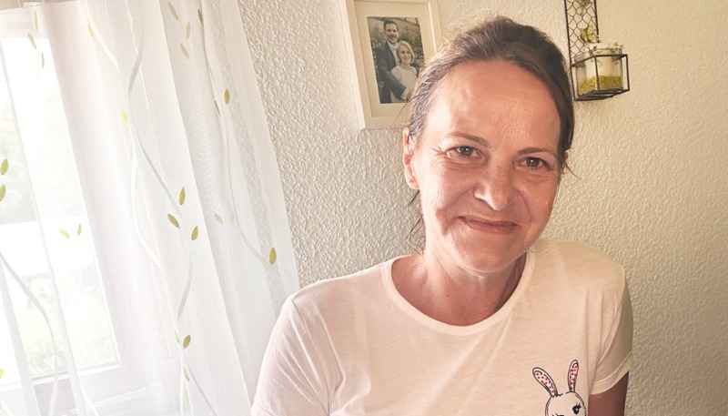 Eine Frau mit braunen Haaren lächelt in einem Innenraum und trägt ein weißes T-Shirt mit einem Hasenmotiv. Im Hintergrund sind ein Fenster mit durchsichtigen Vorhängen und ein gerahmtes Foto zu sehen.