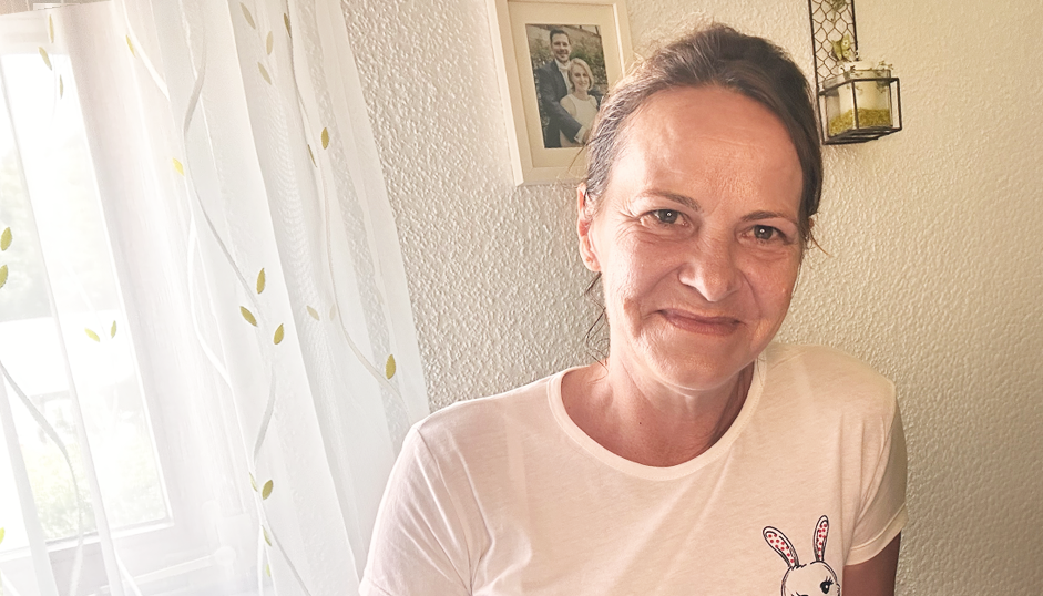 Eine Frau mit braunem Haar lächelt in die Kamera. Sie trägt ein weißes T-Shirt mit einem Hasenmotiv. Sie befindet sich in einem Innenraum, in der Nähe eines Fensters mit durchsichtigen Vorhängen und einem gerahmten Foto an der Wand hinter ihr.