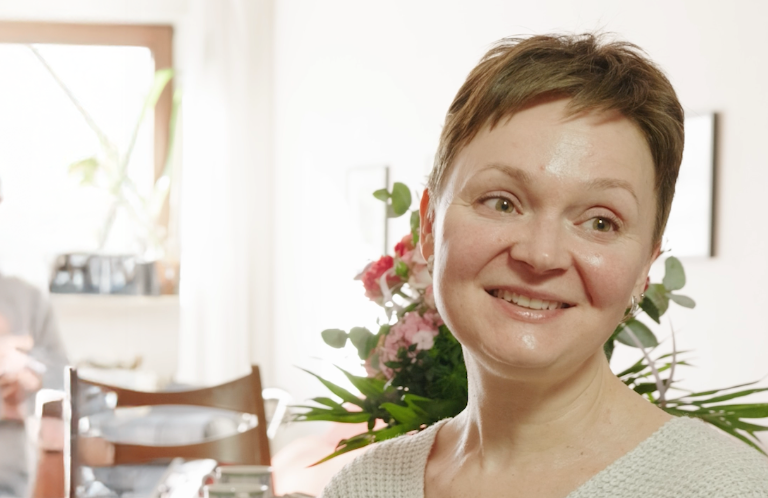 Eine Frau mit kurzen Haaren lächelt sanft, während sie in einem Haus sitzt, mit Blumen und Bücherregalen im Hintergrund. Die Szene ist warm beleuchtet und schafft eine gemütliche und einladende Atmosphäre.
