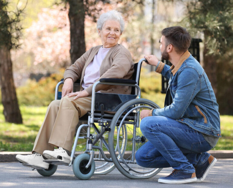 Ein jüngerer Mann kniet neben einer älteren Frau im Rollstuhl im Freien. Beide lächeln sich an. Umgeben von Bäumen und Sonnenlicht genießen sie einen warmen Tag – ein Beispiel dafür, wie Pflegehilfsmittel Unabhängigkeit und Verbundenheit fördern.