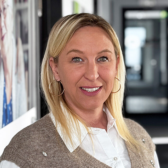 Eine Frau mit langen blonden Haaren, die große Ohrringe, eine weiße Bluse und eine hellbraune Pulloverweste trägt, lächelt, während sie in einem modernen Flur mit verschwommenem Hintergrund und Porträts an der Wand steht.