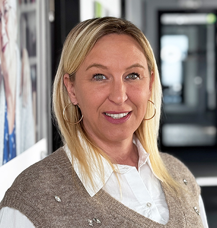 Eine Frau mit langen blonden Haaren, die große Ohrringe, eine weiße Bluse und eine hellbraune Pulloverweste trägt, lächelt, während sie in einem modernen Flur mit verschwommenem Hintergrund und Porträts an der Wand steht.