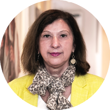Sabine Bernasek, mit schulterlangem braunem Haar, trägt einen gelben Blazer, ein weißes Oberteil, einen Schal mit Animal-Print und große runde Ohrringe, steht drinnen vor einem unscharfen Hintergrund.