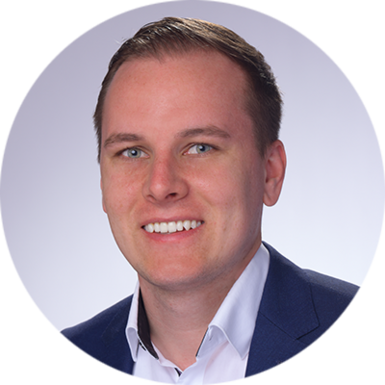 Simon Gsell, ein Mann mit kurzen hellbraunen Haaren, der ein weißes Hemd mit Kragen und einen dunkelblauen Blazer trägt, lächelt vor einem hellgrauen Hintergrund.