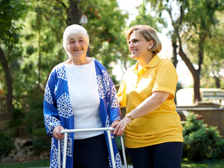 Eine lächelnde ältere Frau mit weißem Haar benutzt im Freien einen Rollator, begleitet von einer fröhlichen Pflegekraft in einem gelben Hemd. Sie stehen in einem sonnigen, grünen Garten mit Bäumen und Pflanzen im Hintergrund.