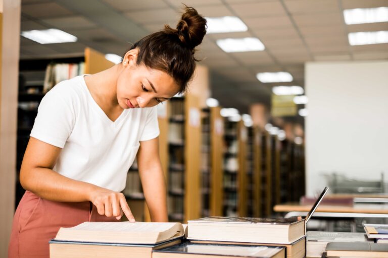 Eine junge Frau steht in einer Bibliothek, liest und zeigt auf eine Seite in einem aufgeschlagenen Ratgeber auf einem Tisch. Vor ihr liegen mehrere Bücherstapel und ein Laptop. Im Hintergrund säumen Bücherregale die Fläche.
