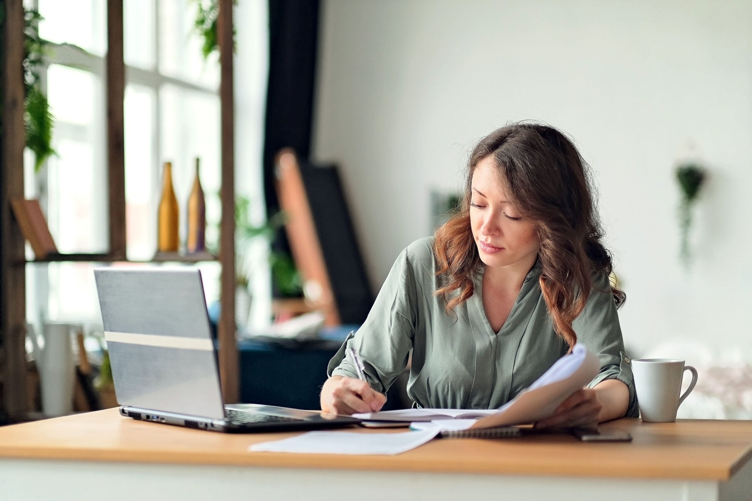 Eine Frau sitzt mit einem Laptop an einem Schreibtisch, hält Papiere in der Hand und schreibt in ein Notizbuch. Sie wirkt konzentriert, liest vielleicht Stellenangebote im Büro. Eine Kaffeetasse steht daneben, und durch das Fenster fällt sanftes Tageslicht.
