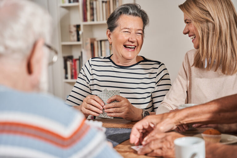 Zwei Frauen lächeln und lachen, während sie mit zwei anderen an einem Tisch Karten spielen und ein fröhliches geselliges Beisammensein in einem gemütlichen, hell erleuchteten Raum genießen – ein Ausdruck der Wärme und Kameradschaft der 24-Stunden-Pflege.