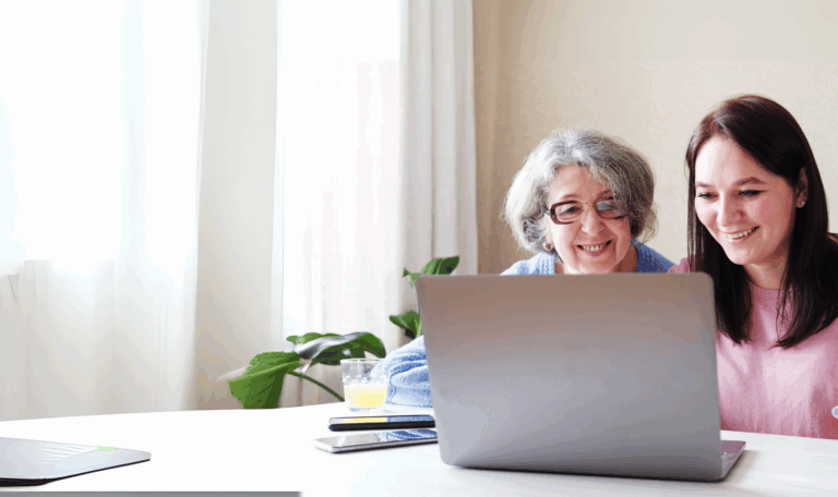 Zwei Frauen, eine ältere mit grauen Haaren und Brille und eine jüngere mit braunen Haaren, sitzen zusammen an einem Tisch und schauen lächelnd auf einen Laptop-Bildschirm, während sie über Osteoporose diskutieren, mit einer Pflanze und einem Glas Saft im Hintergrund.