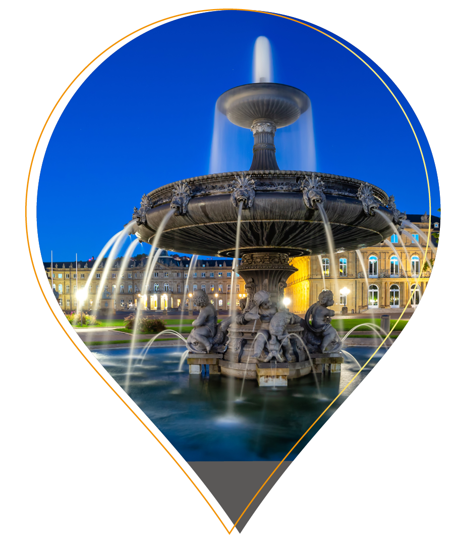Ein großer, verschnörkelter, nachts beleuchteter Springbrunnen mit Wasserläufen und Statuen vor einem historischen Gebäude unter einem tiefblauen Himmel, eingerahmt von einer Landkartennadel auf weißem Hintergrund - ein von Bernd Geisel inspiriertes Kunstwerk.