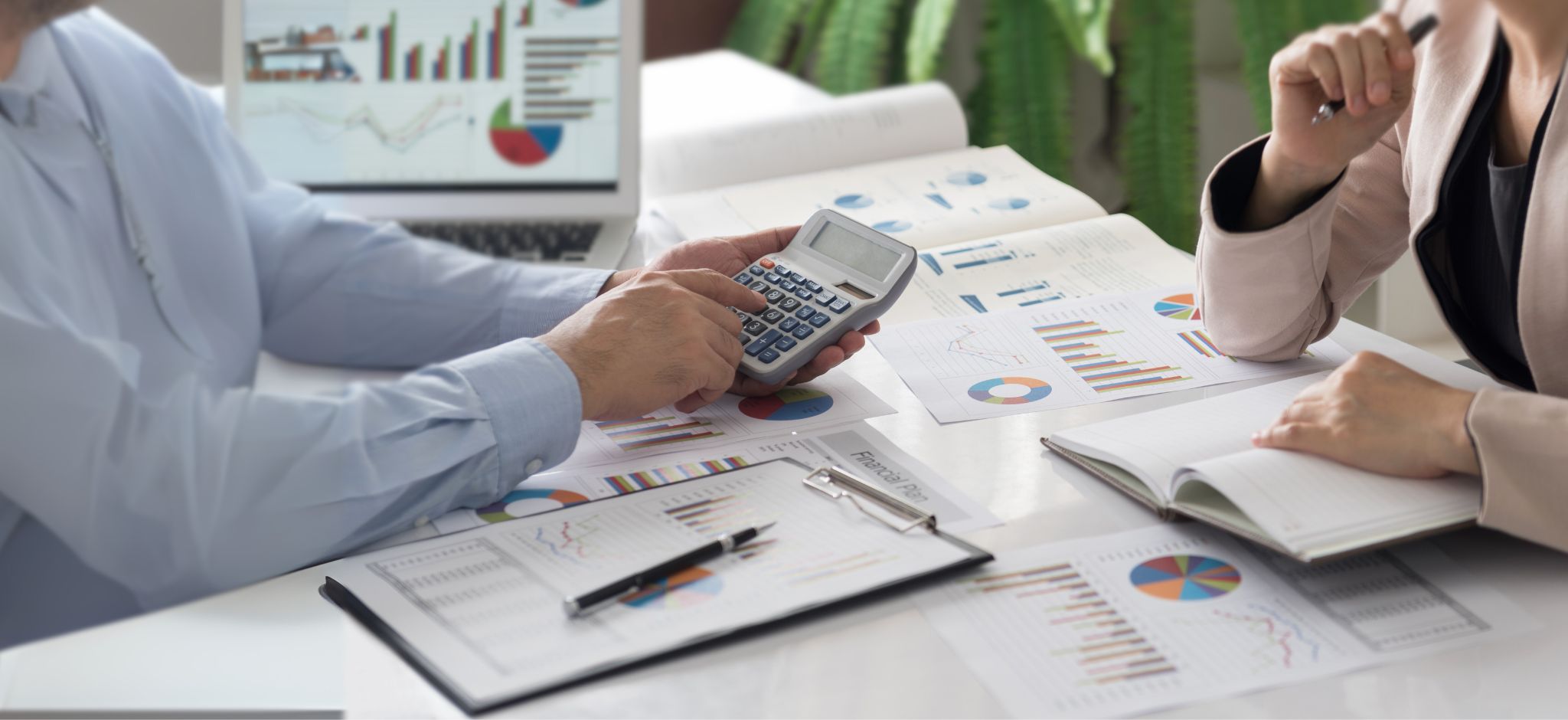 Zwei Personen sitzen an einem mit Finanztabellen und -grafiken übersäten Schreibtisch, die eine hält einen Taschenrechner, die andere ein aufgeschlagenes Notizbuch - so sieht der Arbeitsalltag einer Finanzbuchhalter:in (m/w/d) in Verwaltung und Büroorganisation aus.