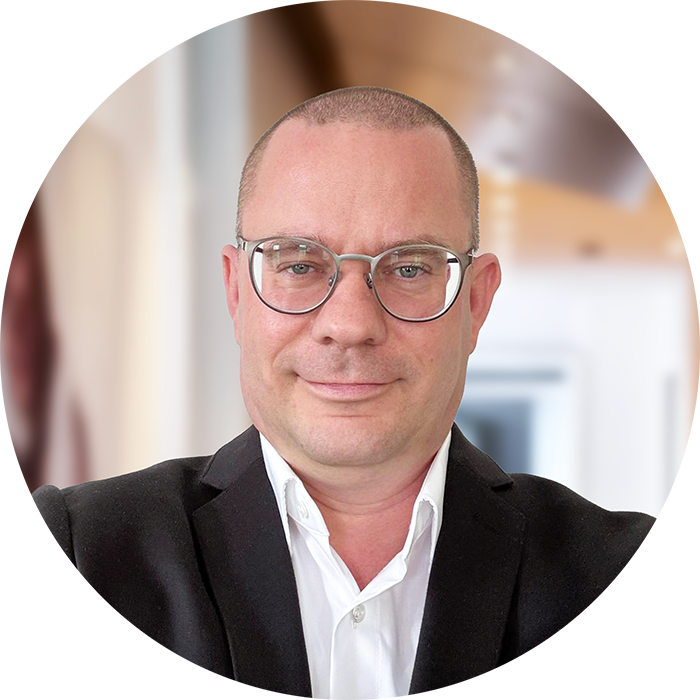 Marcel Lämmel, der eine Brille und eine schwarze Anzugsjacke über einem weißen Hemd trägt, lächelt leicht in die Kamera. Der sanft verschwommene Hintergrund suggeriert eine Büro- oder Wohnumgebung.
