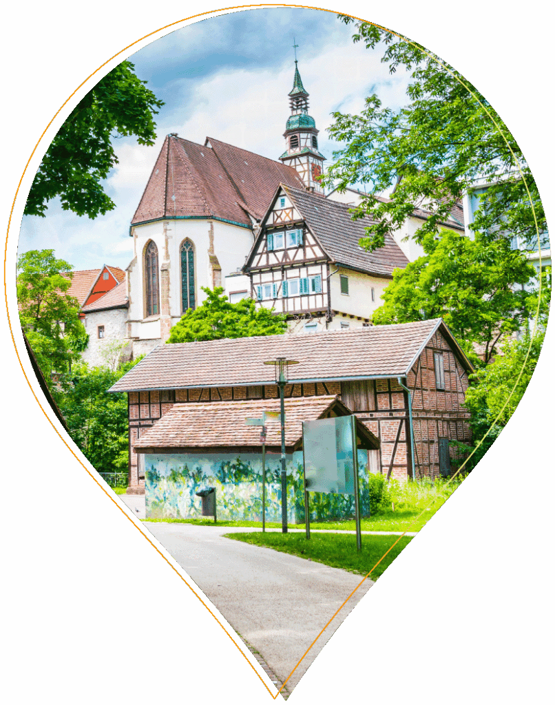 Eine malerische Ansicht einer traditionellen europäischen Kirche mit einem hohen Kirchturm, umgeben von üppig grünen Bäumen und malerischen Fachwerkgebäuden, aufgenommen an einem sonnigen Tag. Das Bild ist von einer Karte in Form einer Stecknadel eingerahmt.