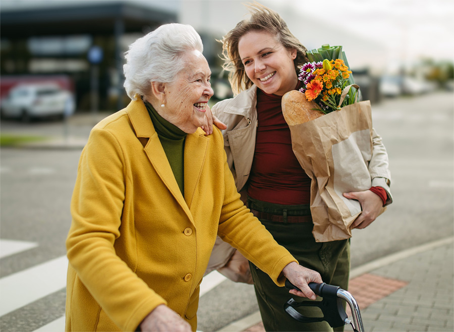 Eine ältere Frau, die einen Rollator benutzt, und ihre Betreuungskraft, die eine Einkaufstasche mit Blumen und Brot trägt, gehen gemeinsam nach draußen, beide lächeln und genießen die Gesellschaft des anderen.