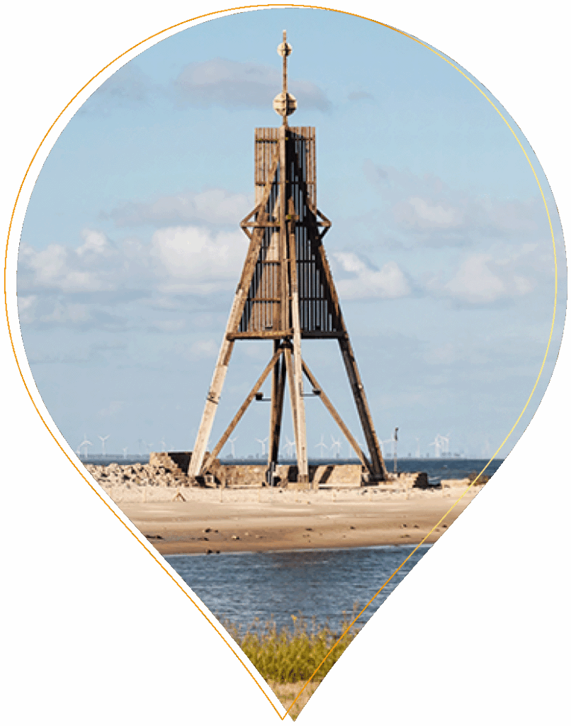 Ein hölzerner Küstenschifffahrtsturm steht an einem Sandstrand in der Nähe des Wassers, und in der Ferne sind bei teilweise bewölktem Himmel Windräder zu sehen.