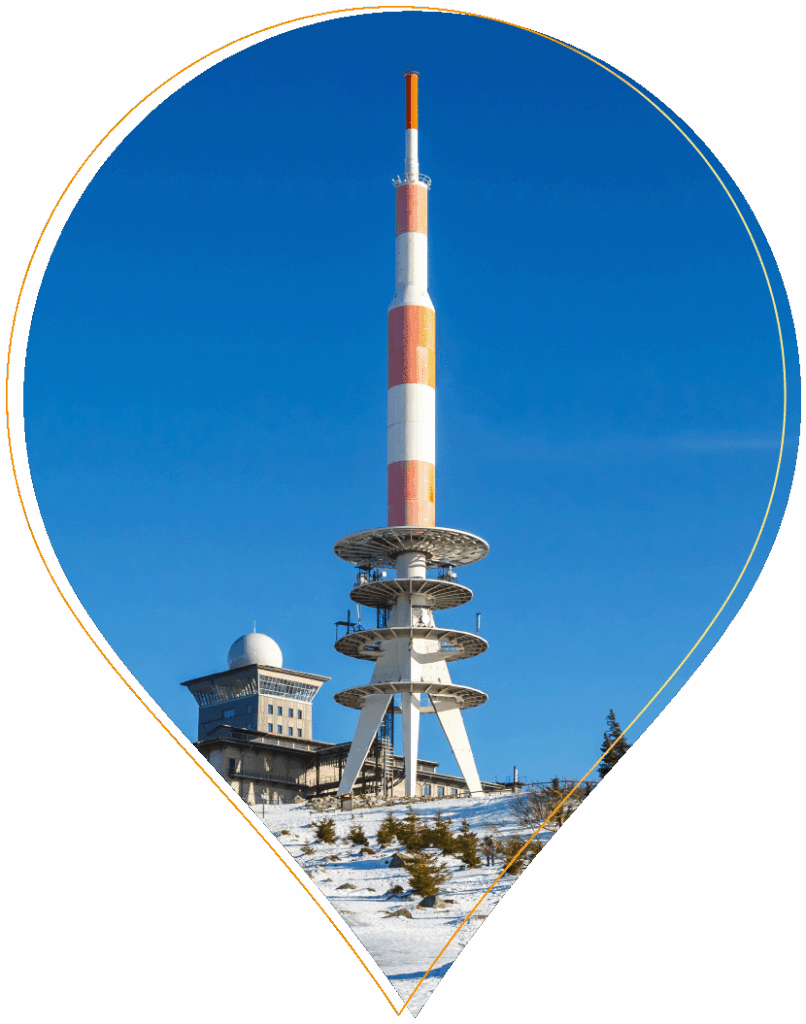 Ein hoher, rot-weißer Antennenturm steht neben einem Gebäude mit Kuppeldach auf einem schneebedeckten Hügel unter einem klaren, blauen Himmel, alles eingerahmt von einer Kartennadelform.