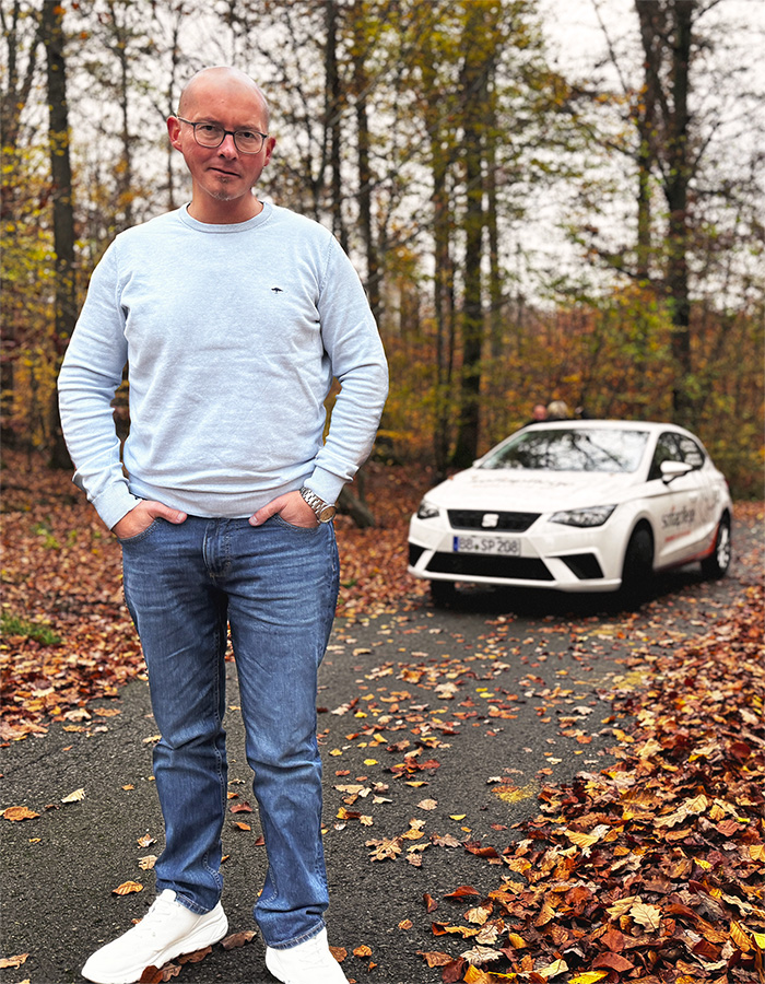 Johannes Vogt, ein Mann in einem hellblauen Pullover und Jeans steht auf einer mit Laub bedeckten Straße in einem Wald, hinter ihm parkt ein weißes Auto. Die Szene ist herbstlich, mit gefallenen Blättern und Bäumen im Hintergrund.