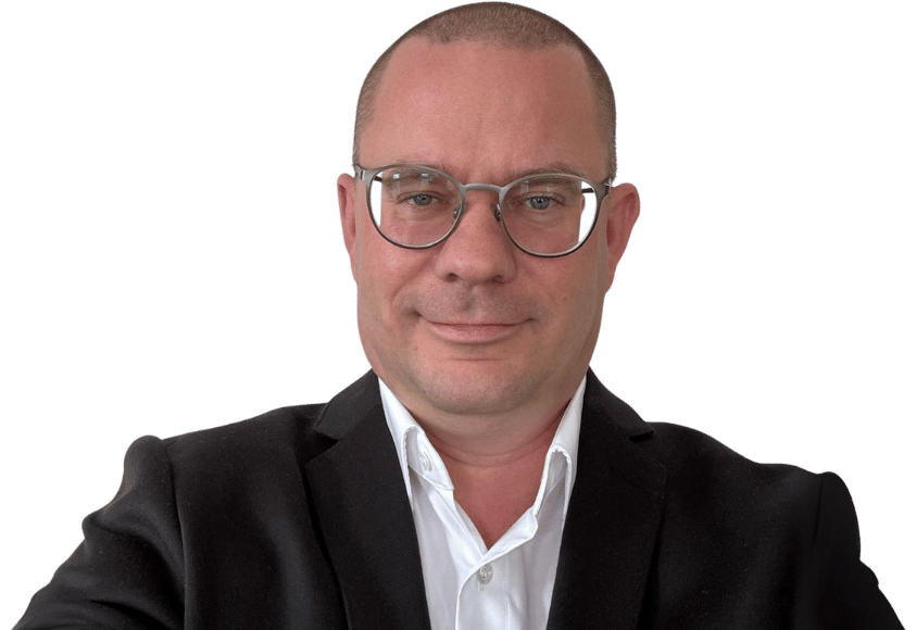 Marcel Lämmel, ein Mann, der eine Brille, ein weißes Hemd und einen schwarzen Blazer trägt, lächelt leicht in die Kamera und posiert vor einem schlichten weißen Hintergrund.