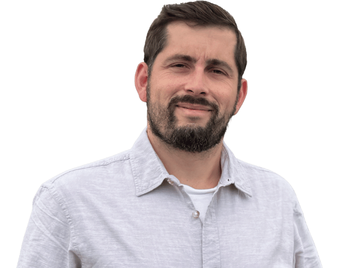 Michael Röder, ein Mann mit kurzen dunklen Haaren und einem Bart steht und lächelt leicht. Er trägt ein hellgraues Button-up-Hemd über einem weißen T-Shirt vor einem schlichten, hellen Hintergrund.