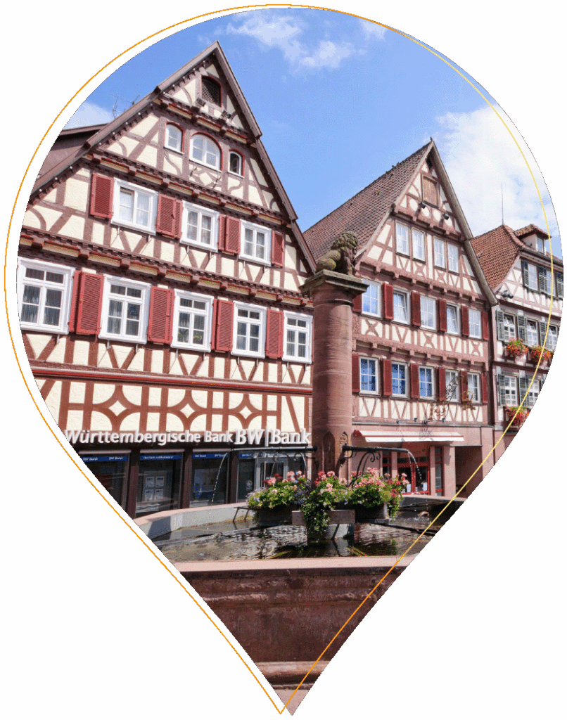 Traditionelle deutsche Fachwerkhäuser mit Blumenkästen, gesehen von der Straßenebene aus an einem sonnigen Tag, innerhalb einer Stecknadelform. Im Erdgeschoss ist ein Schild der Württembergischen Bank zu sehen.