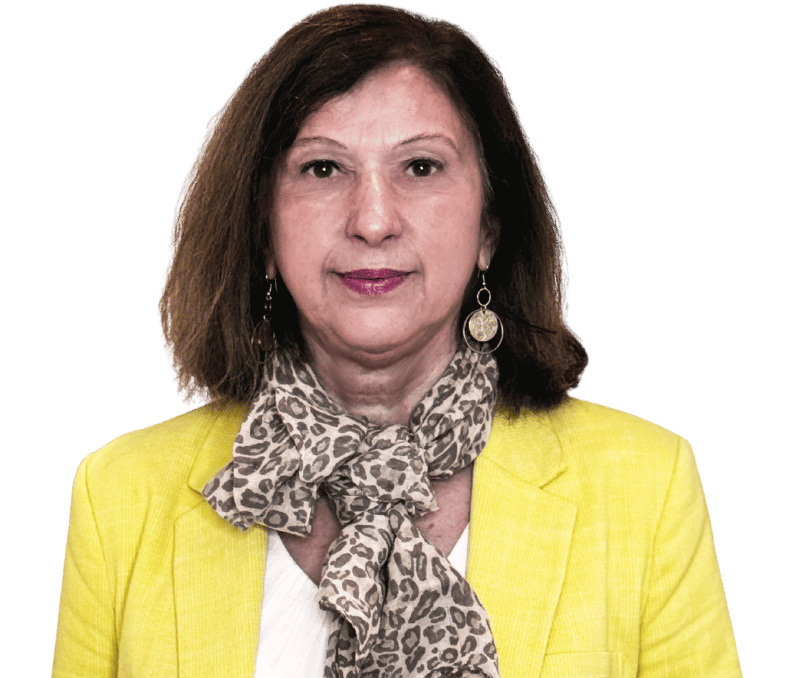 Sabine Bernasek, eine Frau mit schulterlangen braunen Haaren trägt einen gelben Blazer, ein weißes Oberteil, einen Schal mit Leopardenmuster und baumelnde Ohrringe und steht vor einem schlichten hellen Hintergrund.