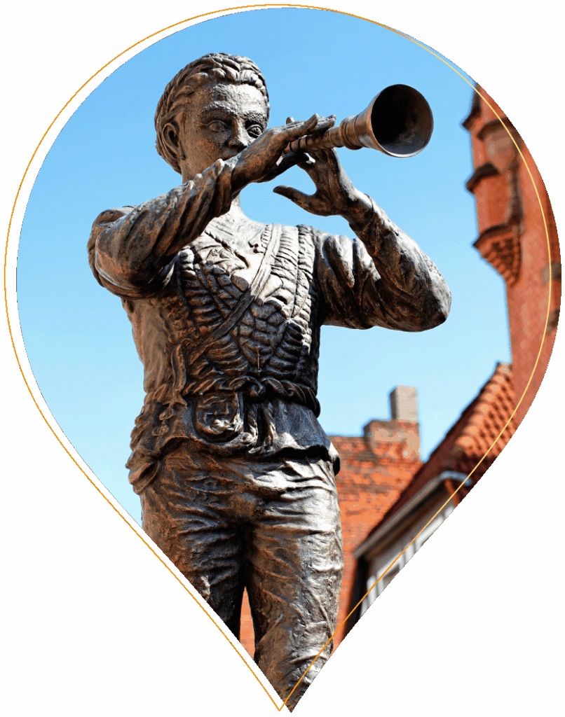 Die Bronzestatue eines Mannes in traditioneller Kleidung, der Pfeife spielt, wurde von Matthias Koepke geschaffen und steht vor einem blauen Himmel und roten Backsteinhäusern.