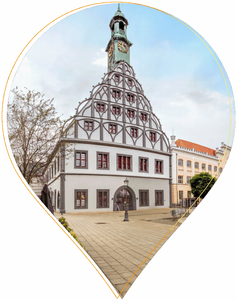 Ein historisches Gebäude mit einem einzigartigen, dekorativen Giebeldach und einem hohen Uhrenturm, auf einem gepflasterten Platz mit Bäumen in der Nähe und angrenzenden klassizistischen Gebäuden, alles unter einem bewölkten Himmel.