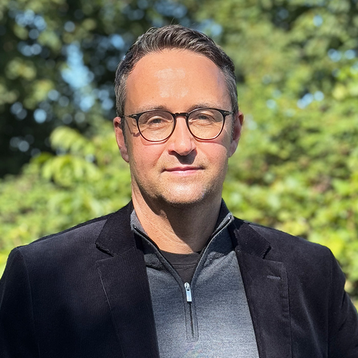 Daniel Waidelich, ein Mann mit kurzen braunen Haaren und Brille, steht im Freien vor grünen Bäumen, trägt einen schwarzen Blazer über einem grauen Reißverschlusshemd und schaut in die Kamera.