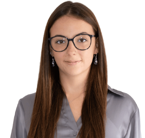 Patricia Wildermuth, eine junge Frau mit langem, glattem braunem Haar und Brille, steht in einer grauen Satinbluse vor einem schlichten, hellen Hintergrund und schaut mit neutralem Blick in die Kamera.