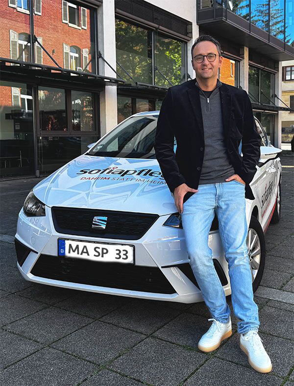 Daniel Waidelich, der eine Brille, einen dunklen Blazer, ein graues Hemd und eine helle Jeans trägt, steht lächelnd an einen weißen SEAT mit Solarfilee-Schriftzug gelehnt auf einer Straße in der Stadt geparkt.