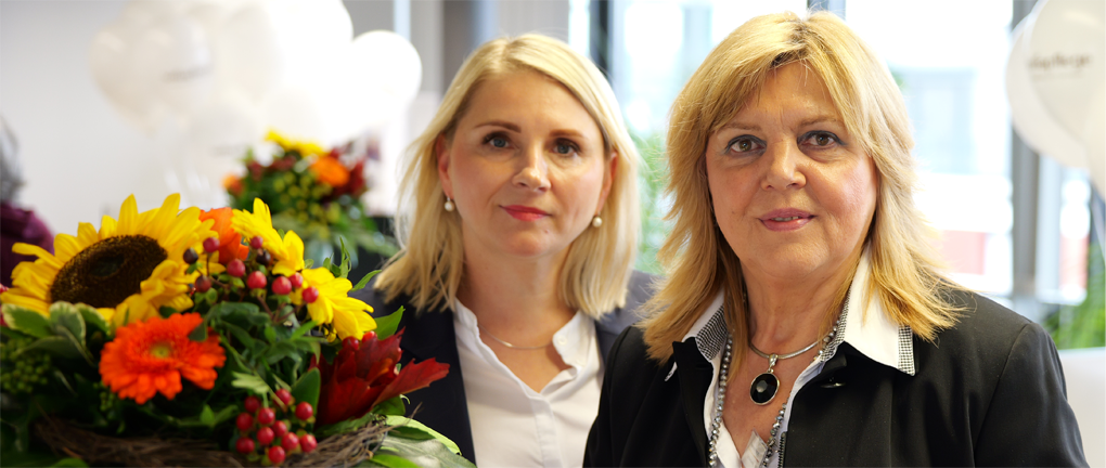 Zwei Frauen mit blondem Haar stehen in einem Haus, beide schauen in die Kamera. Eine Frau hält einen Strauß mit Sonnenblumen und anderen bunten Blumen in der Hand. Der Hintergrund ist sanft beleuchtet, Ballons und Fenster sind zu sehen.