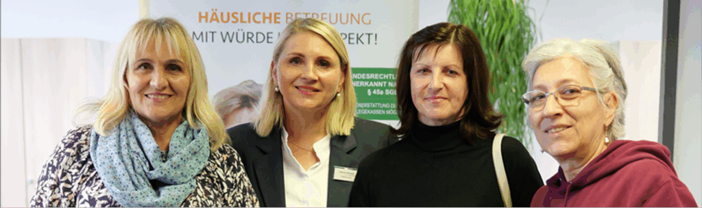 Vier Frauen stehen eng beieinander und lächeln in die Kamera. Hinter ihnen ist ein Transparent mit deutschem Text und einer grünen Pflanze zu sehen. Die Frauen sind in Freizeit- und Geschäftskleidung gekleidet.