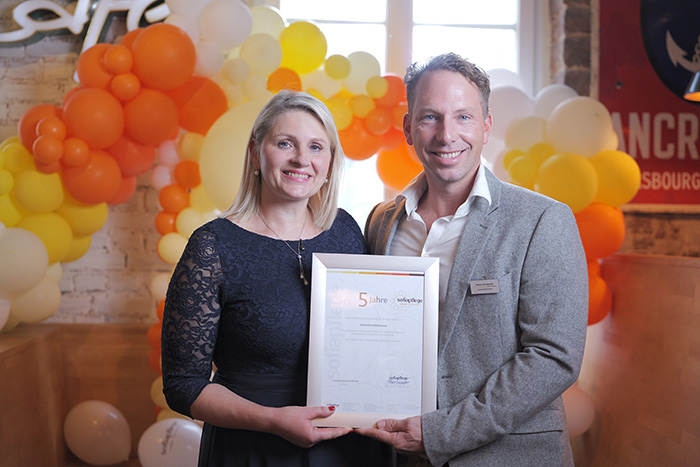 Eine lächelnde Frau und ein lächelnder Mann stehen zusammen in einem Innenraum und halten eine gerahmte Urkunde. Hinter ihnen ist ein dekorativer Bogen aus weißen, gelben und orangefarbenen Luftballons zu sehen, während sie in einem feierlichen Rahmen das Jubiläum - 5 Jahre Sofiapflege Karlsruhe feiern.