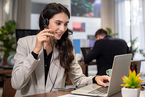 Eine Frau, die ein Headset trägt, lächelt, während sie an einem Büroschreibtisch an einem Laptop arbeitet und möglicherweise während eines Video- oder Telefongesprächs über Stellenangebote diskutiert. Im Hintergrund arbeitet eine weitere Person, und eine kleine Pflanze unterstreicht die Szene.