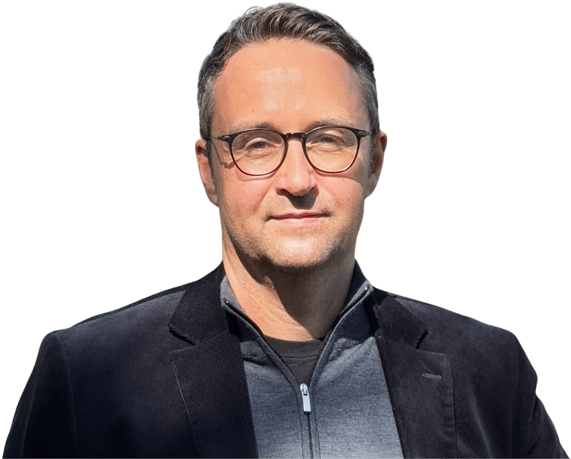 Daniel Waidelich, ein Mann mit kurzen dunklen Haaren und Brille, trägt einen schwarzen Blazer über einem Pullover mit Reißverschluss und steht vor einem schlichten weißen Hintergrund.