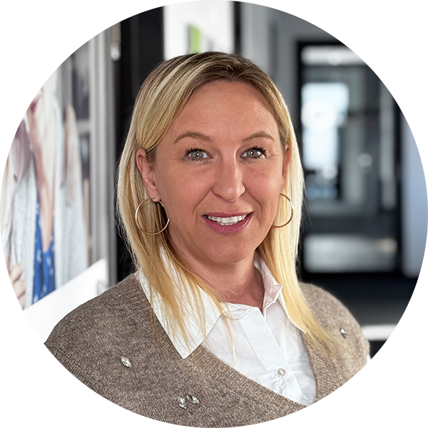 Eine Frau mit glattem blondem Haar, die große Ohrringe, ein weißes Hemd mit Kragen und einen beigen Pullover trägt, lächelt, während sie in einem gut beleuchteten Hausflur steht.