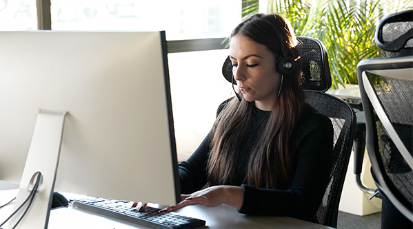Eine Frau mit Headset sitzt an einem Schreibtisch, tippt auf einer Tastatur und blickt auf einen großen Computermonitor in einem hellen Büro mit Grünpflanzen, die als 24-Stunden-Betreuungskraft Unterstützung bieten.