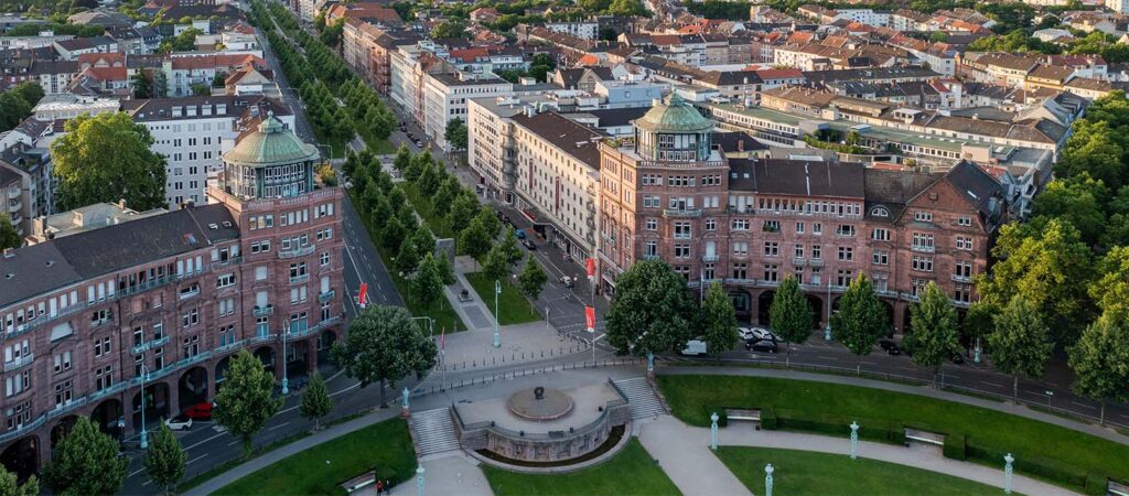 Luftaufnahme eines städtischen Platzes in Mannheim mit einem runden Brunnen, umgeben von grünen Rasenflächen, Bäumen und historischen roten Backsteingebäuden mit Türmen im Herzen der Stadtlandschaft von Mannheim bei der Sofiapflege Metropolregion Rhein-Neckar.