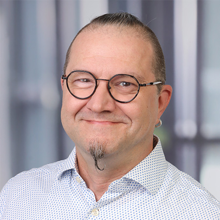 Matthias Wöhr ein lächelnder Mann mit Brille, einem kurzen Bart und einem Kinnbart - Matthias Wöhr - trägt ein weißes Hemd mit kleinen blauen Punkten. Er hat helle Haut, einen Ohrring, sein Haar ist hochgesteckt, und er steht vor einem verschwommenen Hintergrund mit vertikalen Linien.