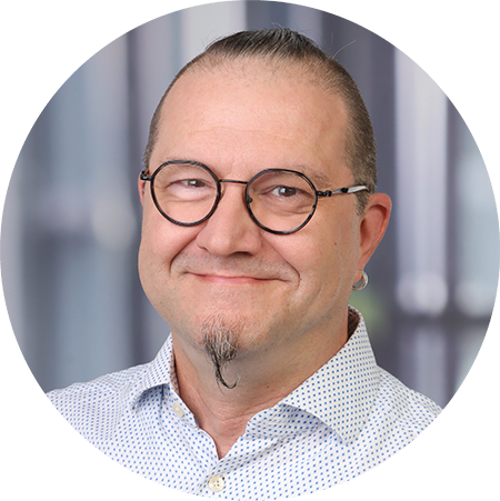 Ein lächelnder Mann mit Brille, einem kurzen Ziegenbart und einem kleinen Ohrring, der ein helles gemustertes Hemd trägt. Der Hintergrund ist mit vertikalen Linien verschwommen.