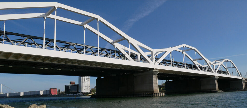 Eine weiße Stahlfachwerkbrücke überspannt einen Fluss mit blauem Himmel darüber. Moderne Gebäude und Wohnungen, Teil der pulsierenden Metropolregion Rhein-Neckar, sind im Hintergrund in der Nähe des Ufers zu sehen.
