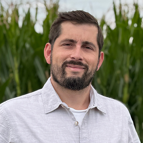 Ein Mann mit kurzen dunklen Haaren und Bart, der ein hellgraues Button-up-Hemd trägt, steht im Freien vor hohen grünen Pflanzen, möglicherweise Mais, und lächelt in die Kamera.